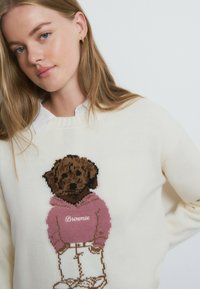 Cremefarbener Pullover mit einem gestrickten Bären, der einen rosa Hoodie und weiße Hose trägt. Das Design zeigt braune Farbtöne für den Bären und detaillierte Gesichtszüge.