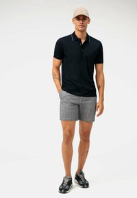 Polo shirt navy con dettagli bianchi, abbinato a pantaloni corti a strisce grigi e bianchi. Il modello indossa un cappello beige e scarpe da ginnastica con fantasia.