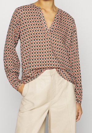 Femme portant un chemisier à manches longues avec un motif losange rouge et vert, rentrée dans un pantalon taille haute crème, une main dans la poche.