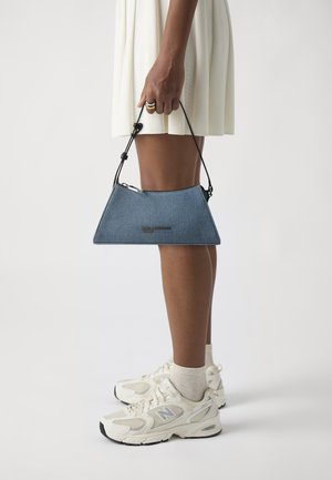 Triangle-shaped denim handbag with a black strap, featuring a zipper and embossed logo. Worn with a white dress and light-colored sneakers.