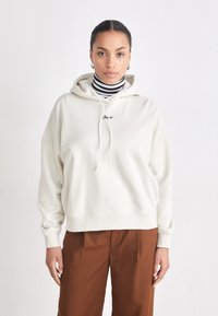 Hanorac oversized crem cu șnur, având un mic logo. Purtați peste un pulover cu gât înalt black și alb cu dungi și pantaloni maro.