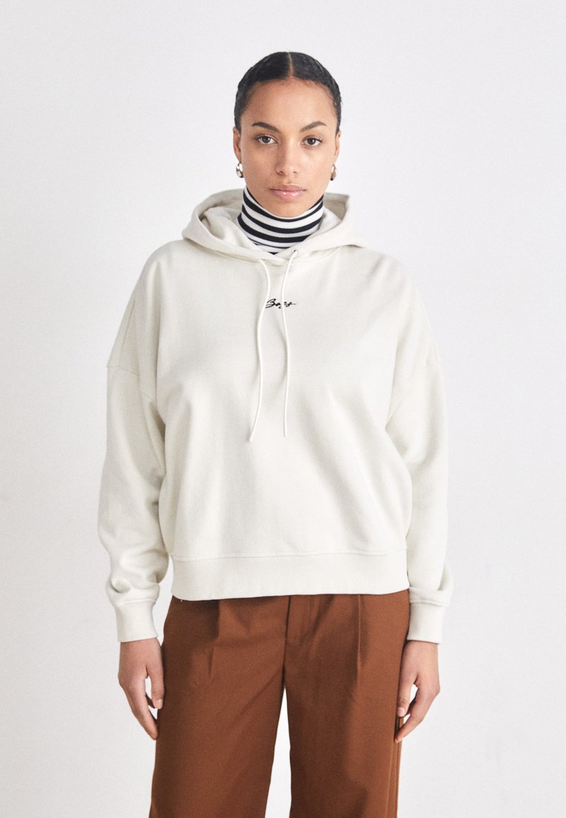 Hanorac oversized crem cu șnur, având un mic logo. Purtați peste un pulover cu gât înalt black și alb cu dungi și pantaloni maro.