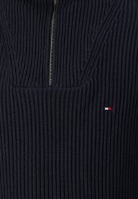 Marineblaues, geripptes Strickpullover mit halbzipper Kragen und einem kleinen rot-weiß-blauen Logo auf der Brust. Strukturierter Stoff mit sichtbaren Nähten.