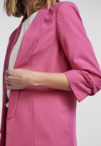 Blazer rose avec une coupe ajustée, tissu léger, col châle et manches retroussées. Détails subtils de coutures le long des coutures.