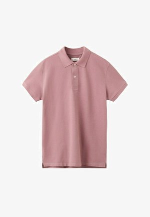 Junger Mann steht mit hinter dem Rücken verschränkten Händen, trägt ein pinkfarbenes Poloshirt, einen hellgrauen Pullover über den Schultern, hellblaue Jeans und beige Sneaker.