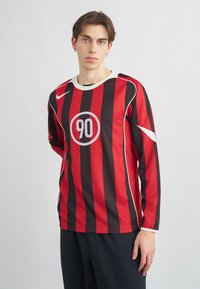 Αθλητική μπλούζα με μακριά μανίκια που διαθέτει ίσες κόκκινες και μαύρες κατακόρυφες ρίγες, στρογγυλό λευκό λογότυπο με τον αριθμό "90" και αντίθετες λευκές λεπτομέρειες.