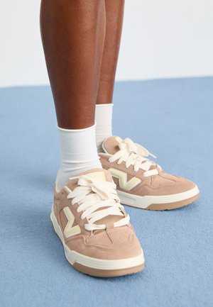 Sapatilhas em camurça castanho claro com detalhes e atacadores brancos, apresentando uma sola espessa e um grande detalhe de logótipo na lateral, usadas com meias brancas.