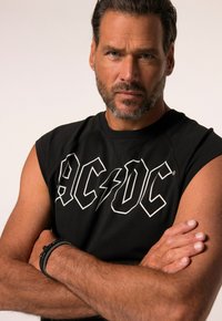 Zwarte mouwloze T-shirt met witte "AC/DC" logo en bliksemschicht. Het model heeft een lichte baard en heeft zijn armen gekruist.