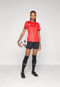 Red short-sleeve soccer jersey with logo, black shorts, black knee-high socks, and black cleats; holding a patterned soccer ball.