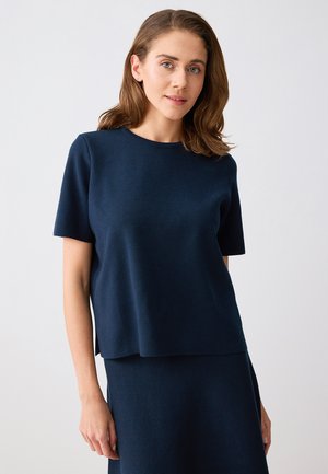 Vrouw met lichtbruin haar, gekleed in een donkerblauw topje met korte mouwen en een bijpassende rok, staand tegen een effen lichte achtergrond.