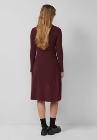 Burgunderfarbenes Strickkleid mit langen Ärmeln, tailliertem Oberteil und ausgestellt geschnittenem Saum, kombiniert mit schwarzen Socken und Schuhen, von hinten betrachtet.