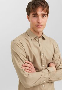 Beige button-up shirt with a collar, featuring long sleeves and a soft, textured fabric. The shirt is worn with arms crossed.