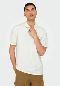 Hvid polo-shirt med en struktureret mønster, korte ærmer og en knaplukning. Båret med khaki shorts; glat stof og klassisk kravedesign.
