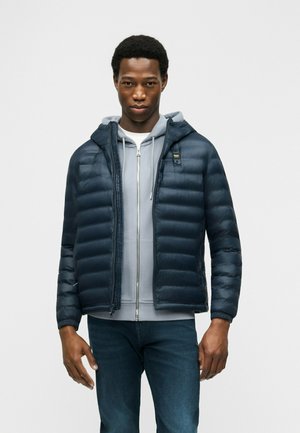 Uomo che indossa una giacca imbottita blu navy sopra una felpa con cappuccio grigia chiara con cerniera e jeans blu scuro, in piedi davanti a uno sfondo semplice.