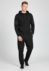 Svart zip-up hoodie med justerbar dragsko, matchande joggerbyxor, båda i slät tyg; har sidofickor och en diskret logodetalj.
