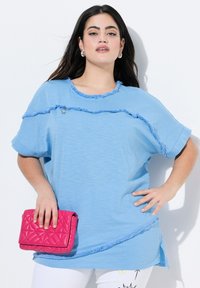 Svetle modré oversized tričko s třepenými okraji a krátkými rukávy, doplněné růžovou prošívanou kabelkou. Hladká textilie, uvolněný střih.