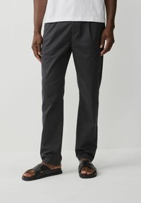 Pantalons en coton gris foncé avec une coupe droite, dotés de plis subtils et d'une texture lisse, associés à des sandales plates noires.
