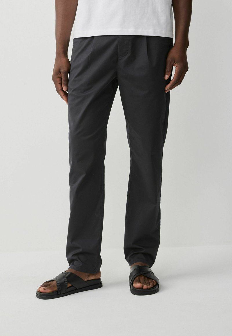 Pantalons en coton gris foncé avec une coupe droite, dotés de plis subtils et d'une texture lisse, associés à des sandales plates noires.