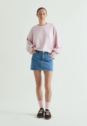 Mujer de pie con las manos en los bolsillos, lleva una sudadera rosa extragrande, falda mini vaquera azul, calcetines rosas y zapatillas negras.