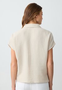 Camicetta beige a maniche corte con finitura testurizzata, caratterizzata da un colletto semplice e una vestibilità rilassata. Il tessuto ha un aspetto leggermente stropicciato.