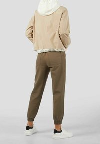 Person iført en beige cropped jakke med hvid hætte, brune bukser med elastisk manchet og hvide sneakers med sort hæl-detalje, stående med hænderne i lommerne.