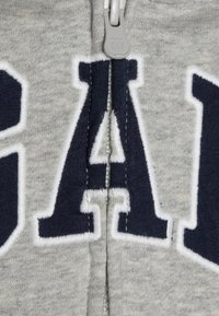 Sudadera con cremallera en color gris, con letras "GAP" bordadas en color azul marino en el frente. Presenta un borde de felpa blanca alrededor de las letras y una cremallera en color gris.