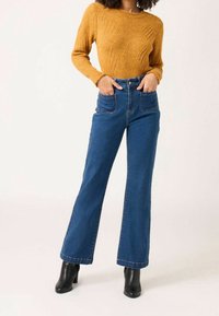 Musztardowy dzianinowy sweter z teksturowanym wzorem diamentu, zestawiony z wysokim stanem niebieskich dzwonowych jeansów i czarnymi botkami na obcasie.