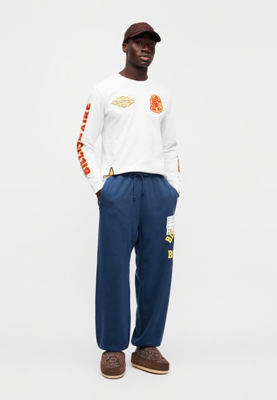 Λευκό πουκάμισο με μακριά μανίκια με κόκκινα και κίτρινα γραφικά, συνδυασμένο με μπλε σκούρο παντελόνι φόρμας με πολύχρωμο κείμενο. Καφέ παπούτσια τύπου slip-on.