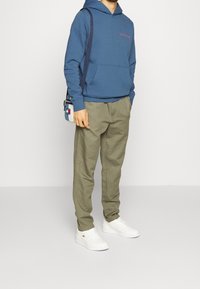Felpa blu con cappuccio, tasca frontale e logo, abbinata a pantaloni tapered di un leggero verde oliva e sneakers bianche. Borsa accessorio piccola indossata a tracolla.