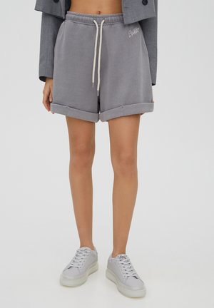Personne portant un short gris à cordon roulé, des baskets blanches et une veste grise courte, debout sur un fond uni.