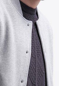 Hellgraue Bomberjacke mit Druckknöpfen, darunter einen grauen gestrickten Pullover mit strukturiertem Muster und einem gerippten Rundhalsausschnitt.