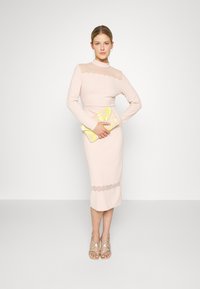 Vestido justo de manga longa em tecido rosa suave, com decote transparente e bainha em forma de onda. A modelo segura uma clutch floral amarela. Sandálias transparentes com tiras.
