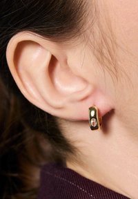 Złota kolczyk w kształcie obręczy o polerowanej powierzchni, z małym owalnym akcentem kamienia szlachetnego, noszony na przekutym uchu, ukazujący gładką teksturę.