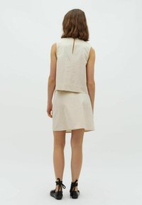 Beige ärmelloses Top mit einem horizontalen Saum und einer Rückenschlitzöffnung, kombiniert mit einem passenden Rock. Schwarze Schnürschuhe runden das Outfit ab.