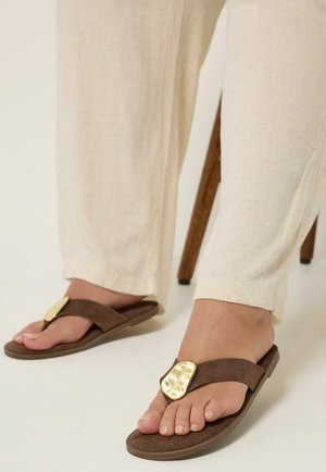 Pieds portant un pantalon en lin beige et des sandales marron avec un ornement ovale doré texturé sur le dessus, debout près d'une patte de chaise en bois.