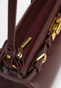 Bordeaux leren handtas met een gestructureerde afwerking, voorzien van gouden hardware, ritssluiting en decoratieve accenten.