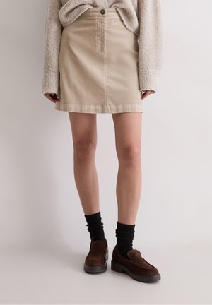 Kobieta w beżowej sztruksowej mini spódniczce, oversize beżowym swetrze, czarnych skarpetkach i brązowych zamszowych mokasynach, stojąca na jednolitym tle.