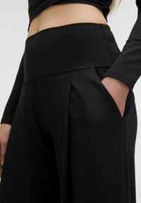 Pantalon noir taille haute avec un détail plissé à l'avant, composé d'un tissu lisse et extensible, doté de poches latérales. Vue rapprochée de la taille à la hanche.