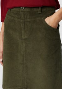 Khaki corduroy-hame, jossa on teksturoitu kangas, etutaskut ja nappikiinnitys, esitettynä käsi taskussa.