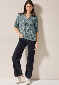 Blusa verde a fantasia con scollo a V e maniche arrotolate, abbinata a jeans a gamba larga blu scuro e sneakers bianche con dettagli verdi.