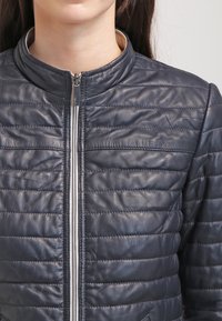 Kobieta w ciemnoniebieskiej pikowanej skórzanej kurtce z przednim zamkiem i stójką, pokazana od szyi do talii.