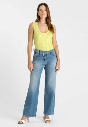 Vrouw draagt een mouwloos geel topje en een high-waist wijd uitlopende blauwe spijkerbroek, staand tegen een witte achtergrond.