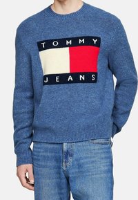 Blå stickad tröja med en djärv grafik med "TOMMY JEANS"-text, vita och röda rektangulära accenter på en marin bakgrund. Ribbad mudd.