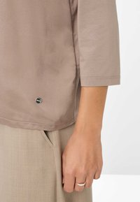 Mano sinistra appoggiata al fianco, indossa un anello d'argento, una camicia a maniche lunghe beige e pantaloni marrone chiaro con cucitura della tasca visibile.