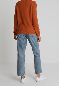 Pull en orange à côtes avec un col rond associé à un jean bleu clair, ample, et des baskets blanches, vu de dos.