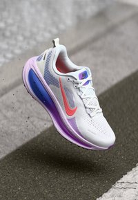 Weißer Laufschuh mit einem atmungsaktiven Mesh-Obermaterial, pinken Akzenten und einer lila Sohle. Hat eine geschwungene Form und gemusterte Details an den Seiten.