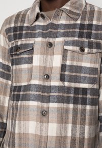 Close-up van een knoopsgatbeige, bruine en grijze geruite shirt met twee knoopsgat borstzakken.