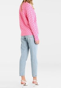 Mallikka seisoo neutraalissa taustassa pinkissä pilkkupaidassa, sinisissä korkeavyötäröisissä farkuissa ja valkoisissa korkokengissä.