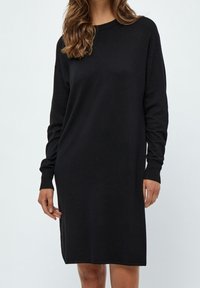 Modella che indossa un abito in maglia nero a maniche lunghe, in piedi contro uno sfondo chiaro, con lunghi capelli che le cadono sulle spalle.