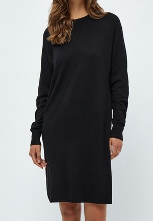 Mannequin portant une robe tricotée noire à manches longues, se tenant contre un fond clair, avec de longs cheveux tombant en cascade.